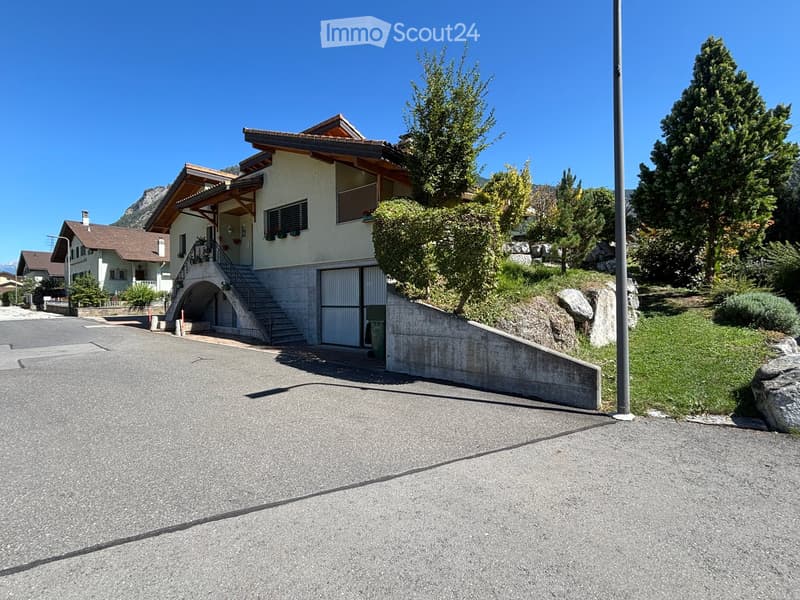 Two-story house with a garage, raised ground floor, and surrounding greenery.