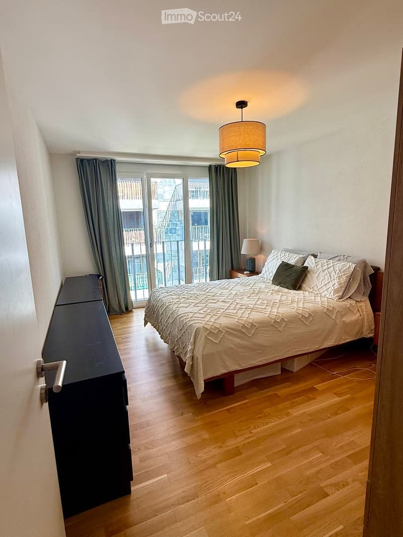 Cozy bedroom with wooden flooring, a large bed with white bedding, a nightstand with a lamp, and a pendant light hanging from the ceiling. The room features large windows with grey curtains and a sliding glass door leading to a balcony.