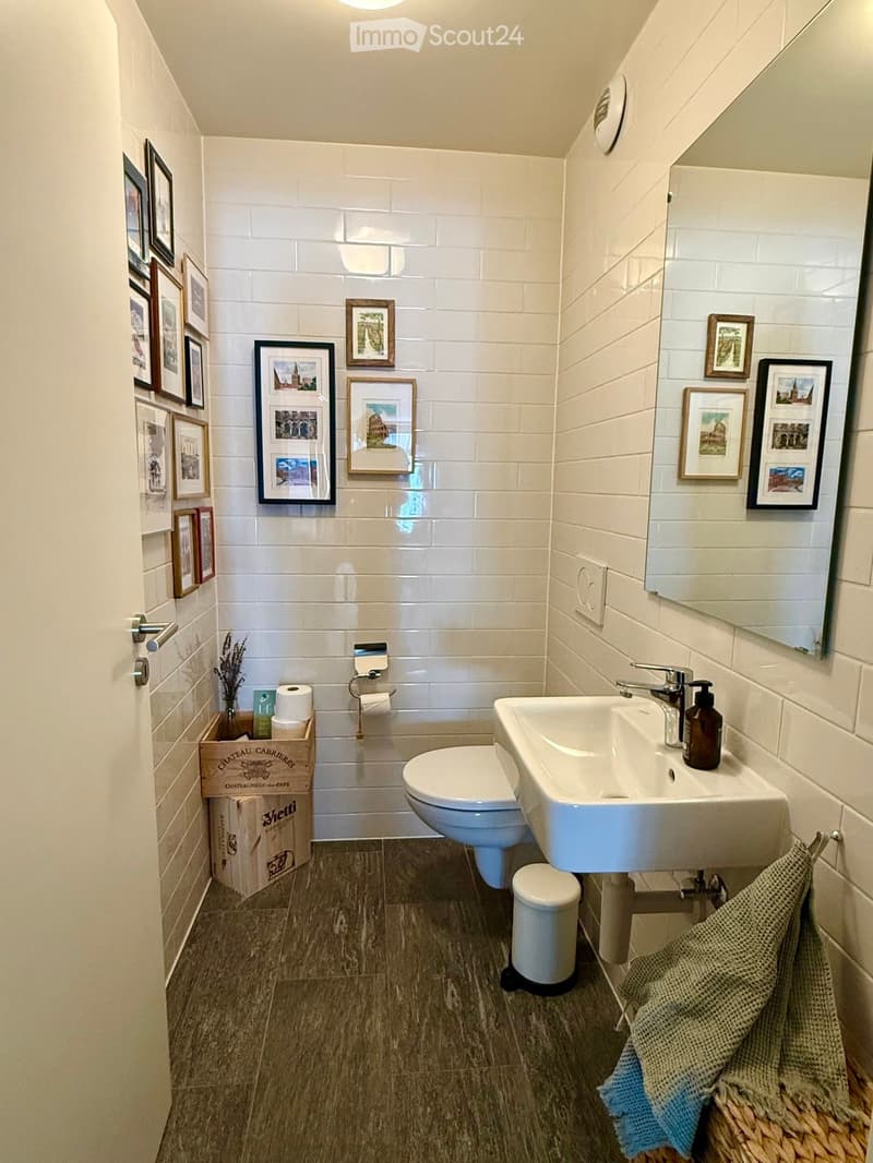 Modern bathroom with white tiles, a toilet, a sink with a mirror, a soap dispenser, a towel, and picture frames on the walls.