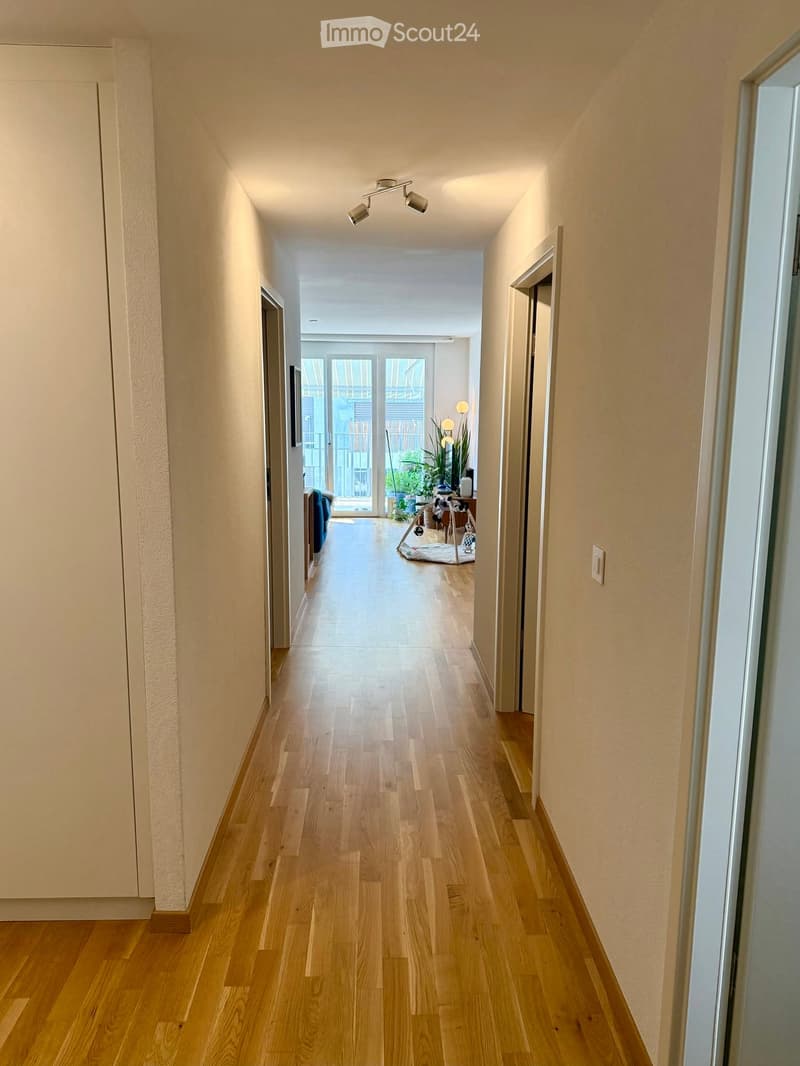Long, narrow hallway with light wooden flooring, white walls, and recessed ceiling lighting. The hallway leads to an open area with a view of a balcony and some indoor plants visible at the end.