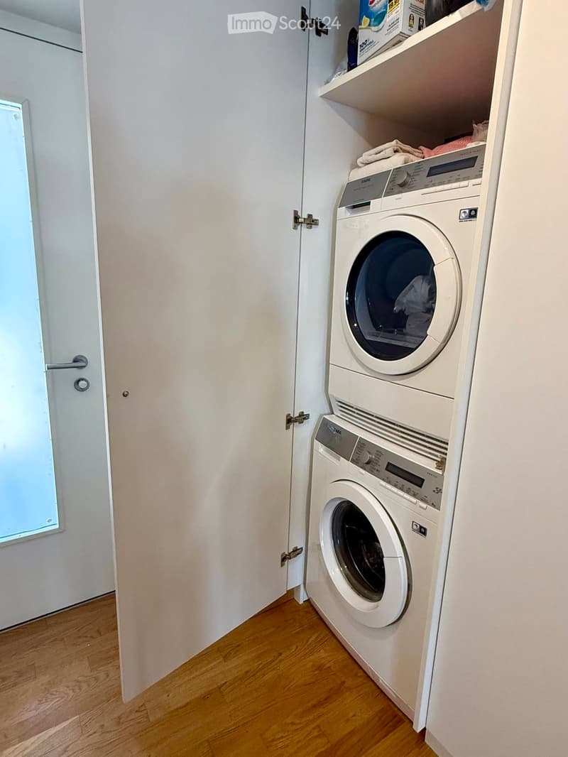 Compact bathroom with a stacked washing machine and dryer unit, wooden flooring, and built-in shelving above the appliances.