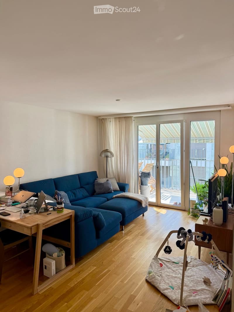 Bright living room with wooden flooring, large sliding glass doors leading to a balcony, blue sectional sofa, wooden side table with laptop, floor lamp, and baby playpen.