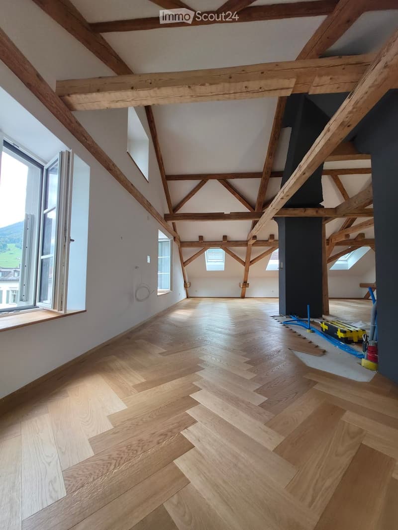 Empty attic space with exposed wooden beams, wooden flooring, and large windows allowing natural light. No furniture or equipment present.