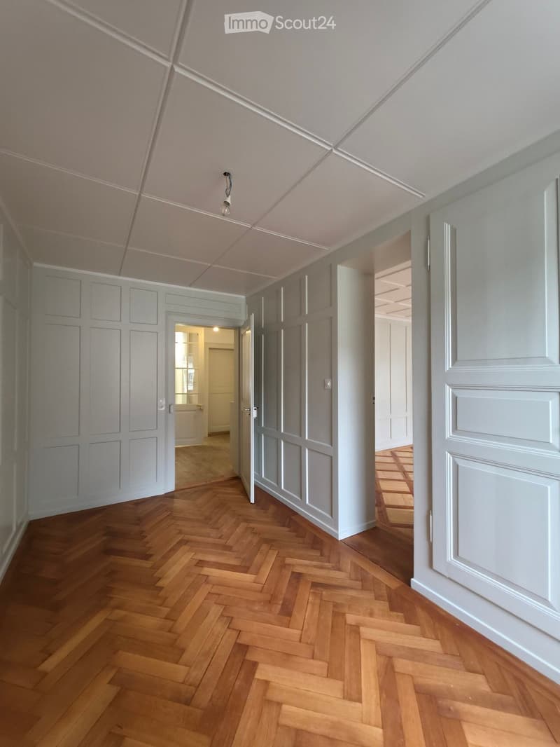 Empty hallway with wooden herringbone flooring, white walls, and several doors leading to other rooms.