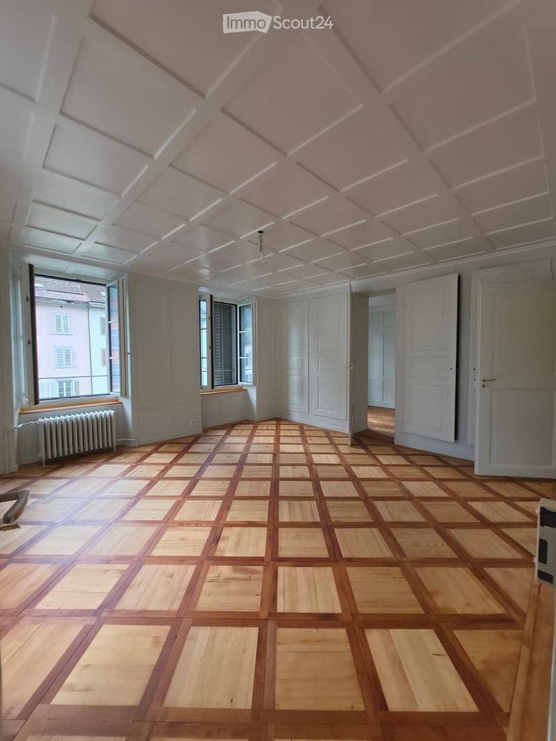 Empty room with wooden parquet flooring, white walls, large windows, and a radiator. The ceiling features a decorative, coffered design with integrated lighting.