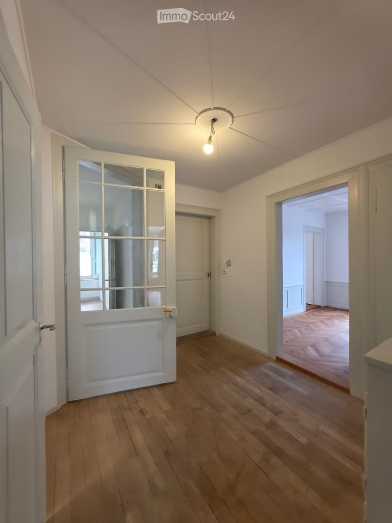 Empty hallway with wooden flooring, white walls, and a ceiling light fixture. Doors lead to adjacent rooms.