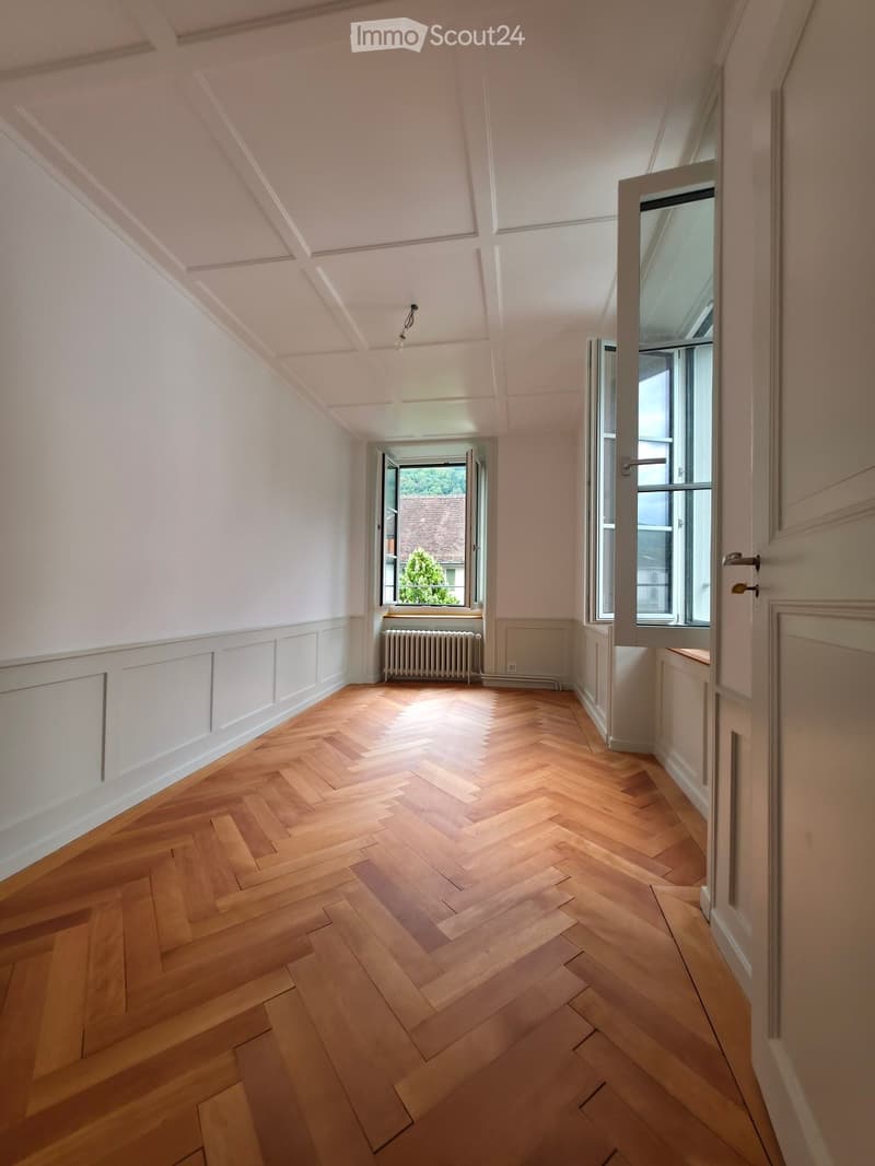 Empty room with wooden herringbone flooring, white walls, and large windows letting in natural light. The room has a radiator and a door leading to another space.