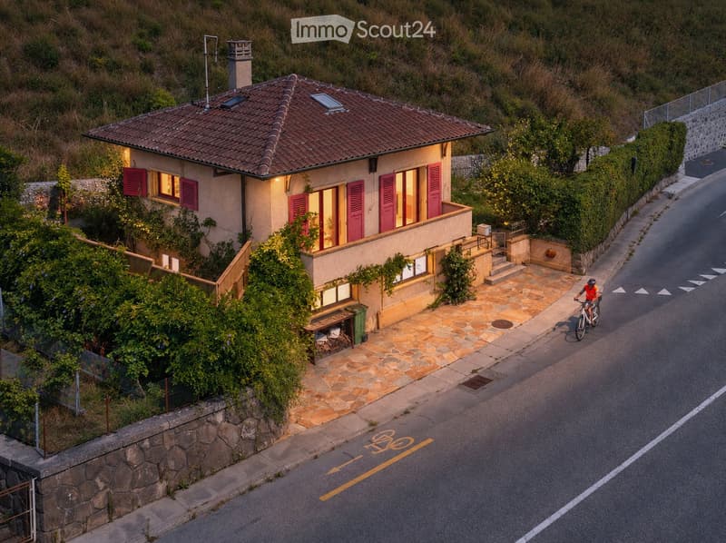 Two-story house with a tiled roof, red shutters, and a stone pathway leading to the entrance. The house is surrounded by greenery and has a small balcony area.