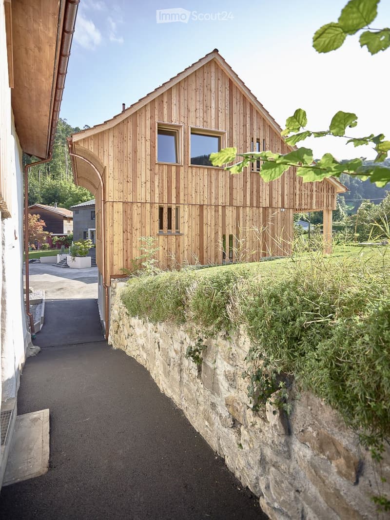 A modern wooden house with a pitched roof and large windows, situated on a raised ground floor with a stone retaining wall and green grass on top.