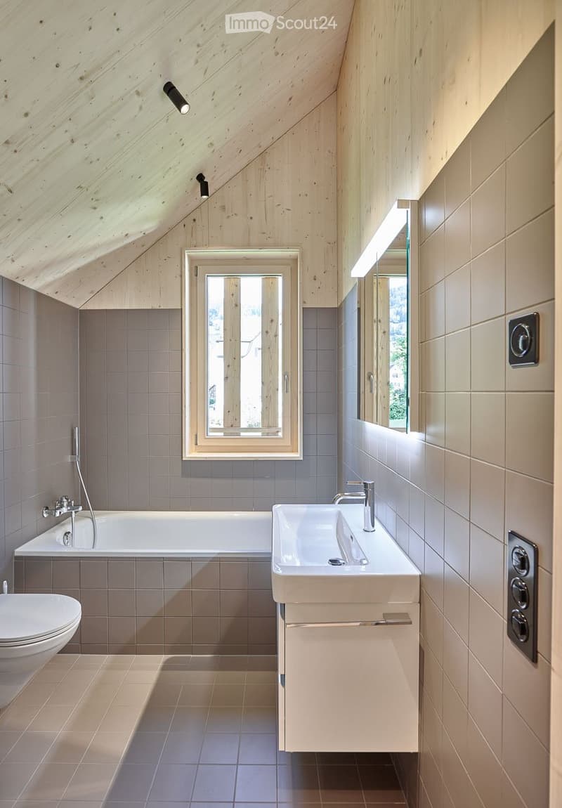 Modern bathroom with wooden ceiling beams, large window, bathtub, toilet, sink, and mirror.