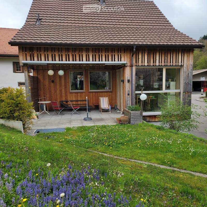 A modern wooden house with a tiled roof, surrounded by a well-maintained garden with colorful flowers and a patio area featuring outdoor furniture.