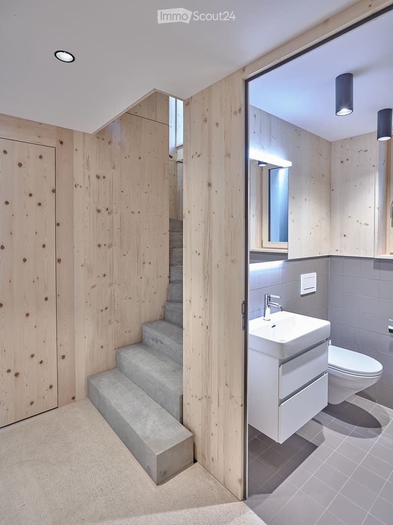 Modern bathroom with wooden walls, a white sink, toilet, and a large mirror. The space features a small staircase leading up, and recessed lighting.