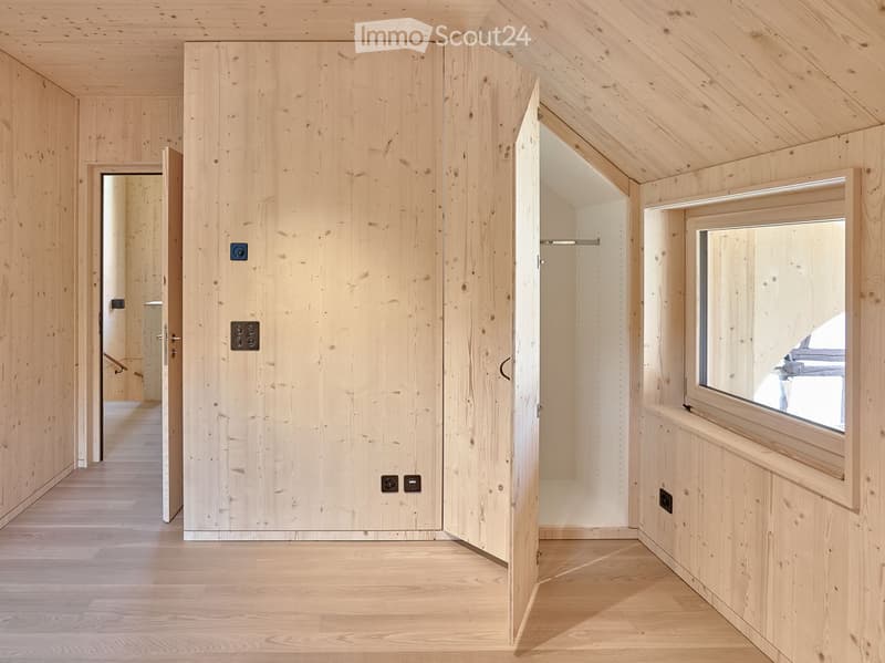 Minimalist wooden hallway with light wood paneling, a closed door, and an open doorway leading to another room. Features a large window on the right side, allowing natural light to enter.