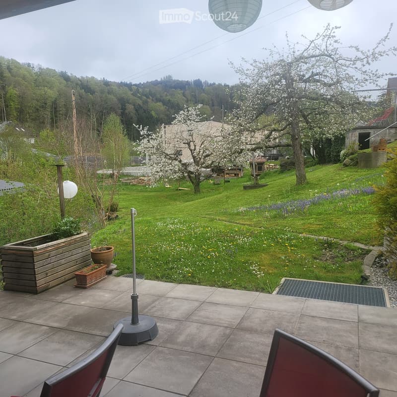 Garden with a patio area, chairs, umbrella, and potted plants, surrounded by a grassy lawn and blooming trees.