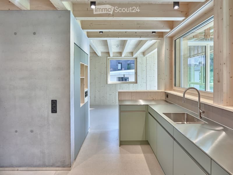 Modern kitchen with light wood cabinetry, stainless steel sink, and a sleek countertop. Exposed wooden beams on the ceiling and large windows provide natural light.