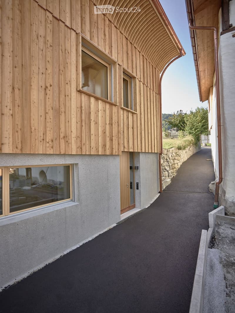 A modern, narrow pathway between two buildings with wooden and concrete facades, featuring large windows and a stone wall on one side.