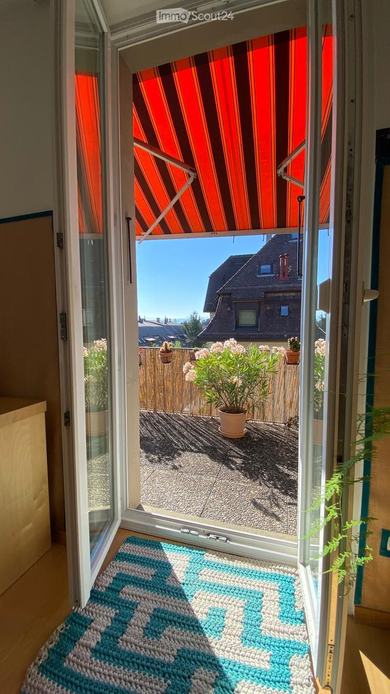 Open sliding glass door leads to a small balcony with a view of the neighborhood, featuring potted plants and a decorative rug inside the doorway.