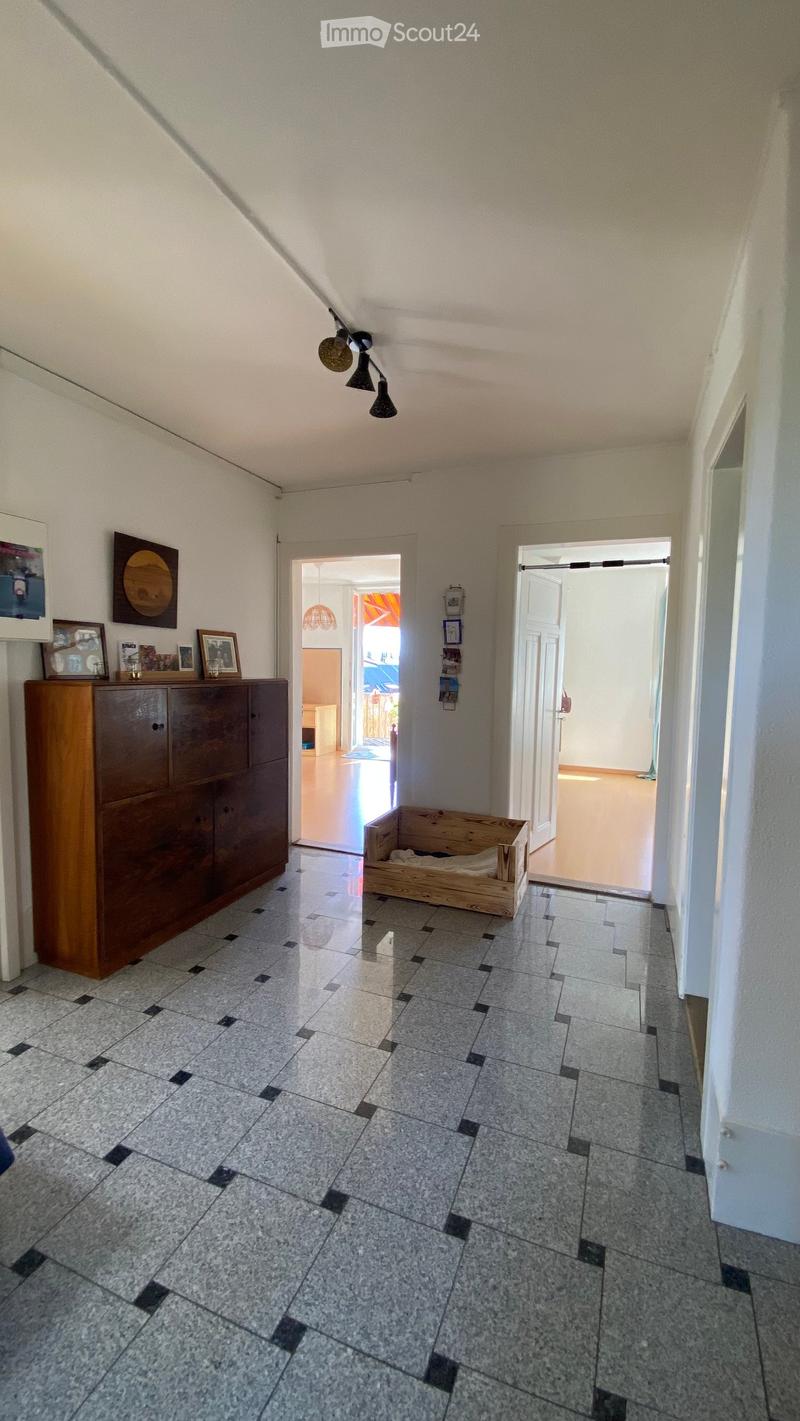 A clean, well-lit hallway with tiled flooring and white walls. Features include a wooden cabinet with framed pictures, a small wooden pet bed, and several doors leading to other rooms.