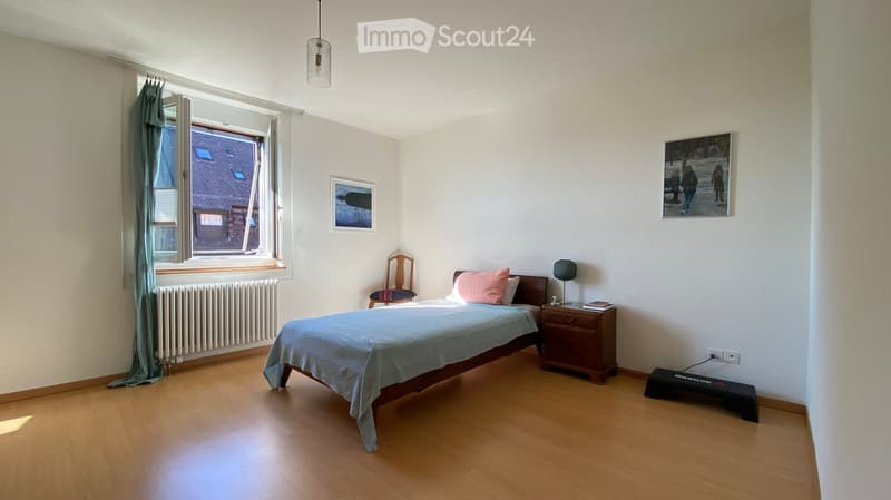 Cozy bedroom with wooden flooring, a bed with blue bedding, a nightstand with a lamp, a wooden chair, and framed pictures on the walls.