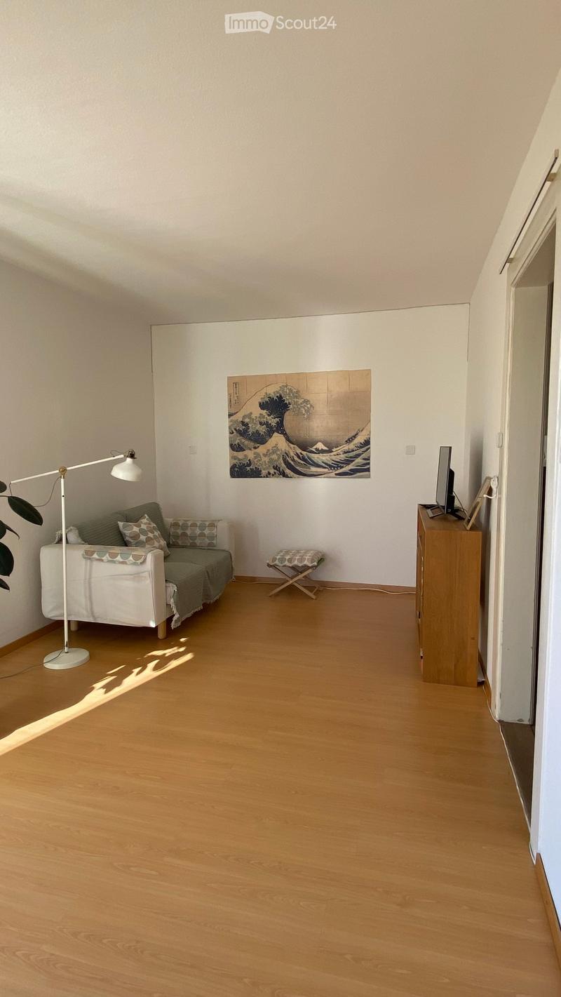 A modern living room with a white sofa, green cushions, a floor lamp, a small wooden stool, a wooden table with a screen, and a large wave-themed picture on the wall.