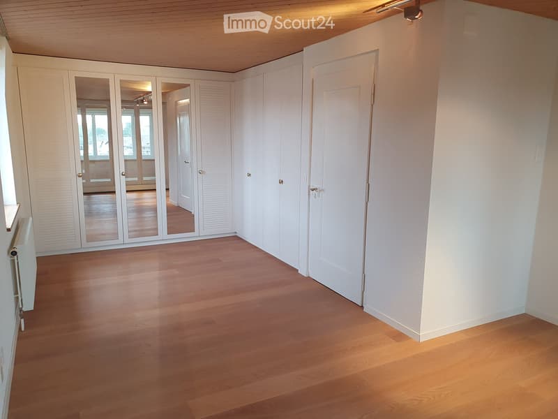 Empty room with light wooden flooring, white walls, and a large mirrored wardrobe unit. The room has a door leading to another space and a radiator on the left side.