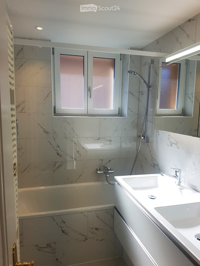 Modern bathroom with marble tiles, a bathtub, a double sink, and a large mirror. The space features sleek chrome fixtures and a frosted glass window above the bathtub.