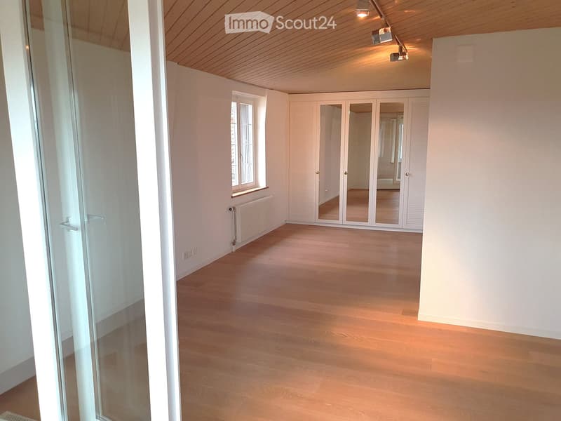 Empty room with light wooden flooring, white walls, large window, mirrored closet, and glass door leading to another space.