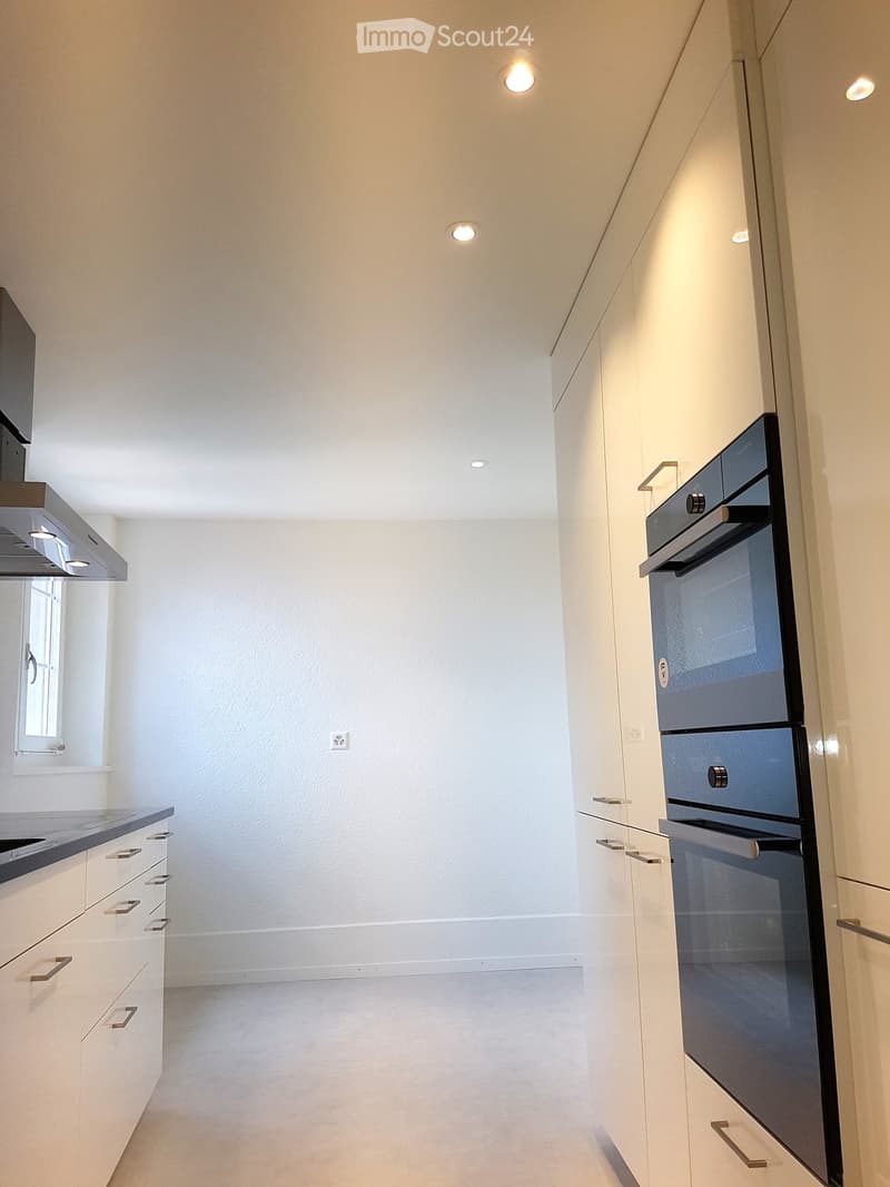 Modern kitchen with white cabinets, stainless steel oven, and range hood. Recessed ceiling lights and a small window provide illumination and ventilation.