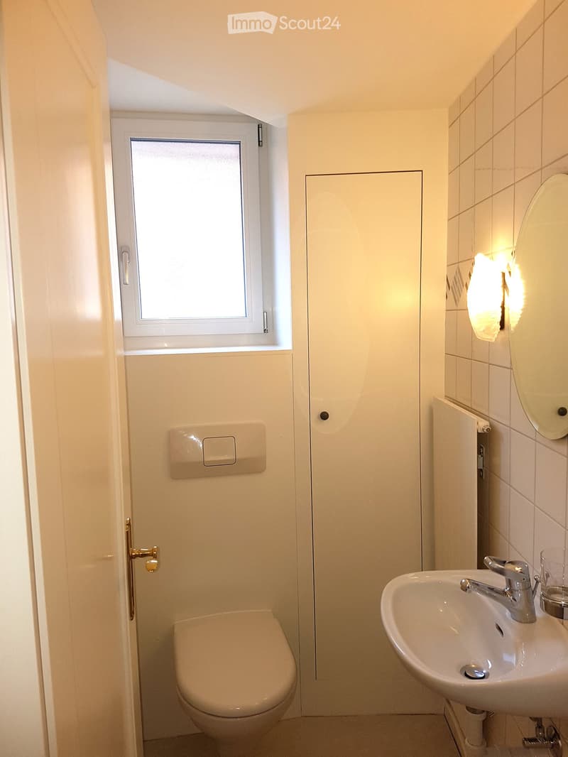 Modern bathroom with white tiles, a toilet, a sink with a chrome faucet, and a mirror above the sink. A small window provides natural light.