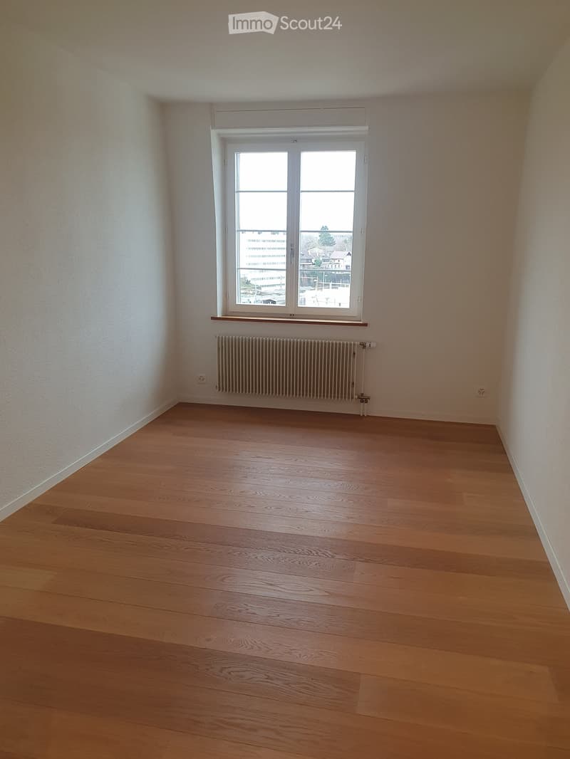 Empty room with wooden flooring, white walls, a window with a radiator below, and a small shelf above the radiator.