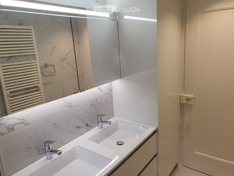 Modern bathroom with a double sink vanity, marble tiles, sleek chrome faucets, and a large mirror with built-in lighting.