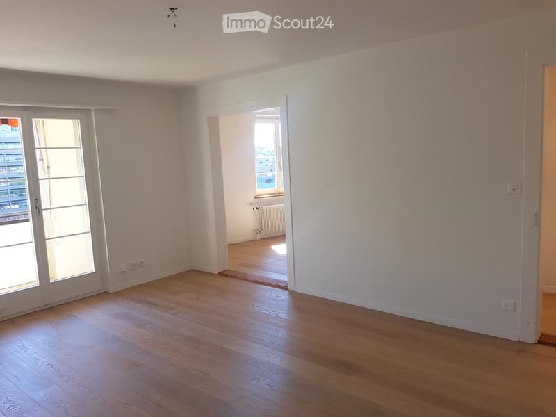 Empty room with light wooden flooring, white walls, and large windows with white frames. The room has a sliding glass door leading to a balcony or outdoor area.