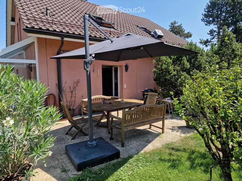 Garden area with a wooden dining table, chairs, and a large umbrella, surrounded by greenery and a house with a tiled roof.