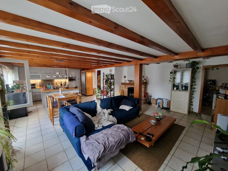 Open-plan living and dining area with exposed wooden beams, a blue sectional sofa, a wooden coffee table, a dining table with chairs, a kitchen area with a refrigerator, oven, and island, and a fireplace.