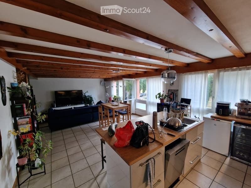 Open-plan living and dining area with a modern kitchen, wooden beams on the ceiling, and large windows. Features a sofa, dining table with chairs, refrigerator, dishwasher, coffee maker, and various kitchen items.