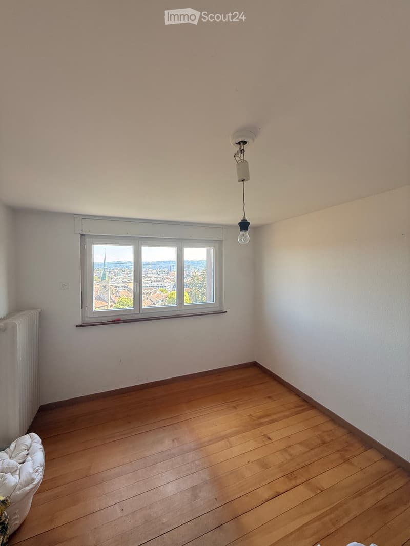 Empty room with wooden flooring, white walls, a single window offering a cityscape view, and a simple light fixture on the ceiling.