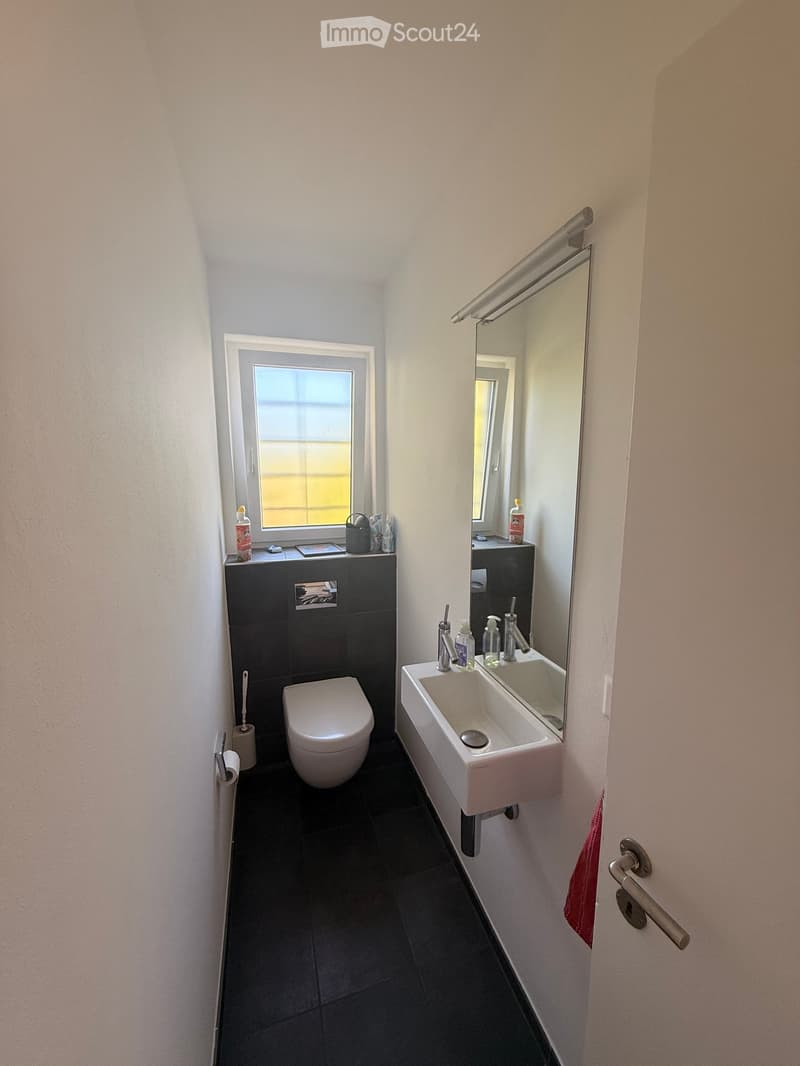 Modern bathroom with white walls, dark tiled floor, toilet, sink, mirror, and window with frosted glass.