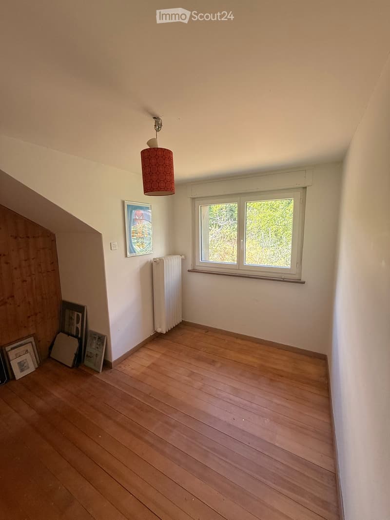 Empty room with wooden flooring, white walls, a window with a view of greenery, a radiator, a hanging lamp, and a few stacked picture frames in the corner.