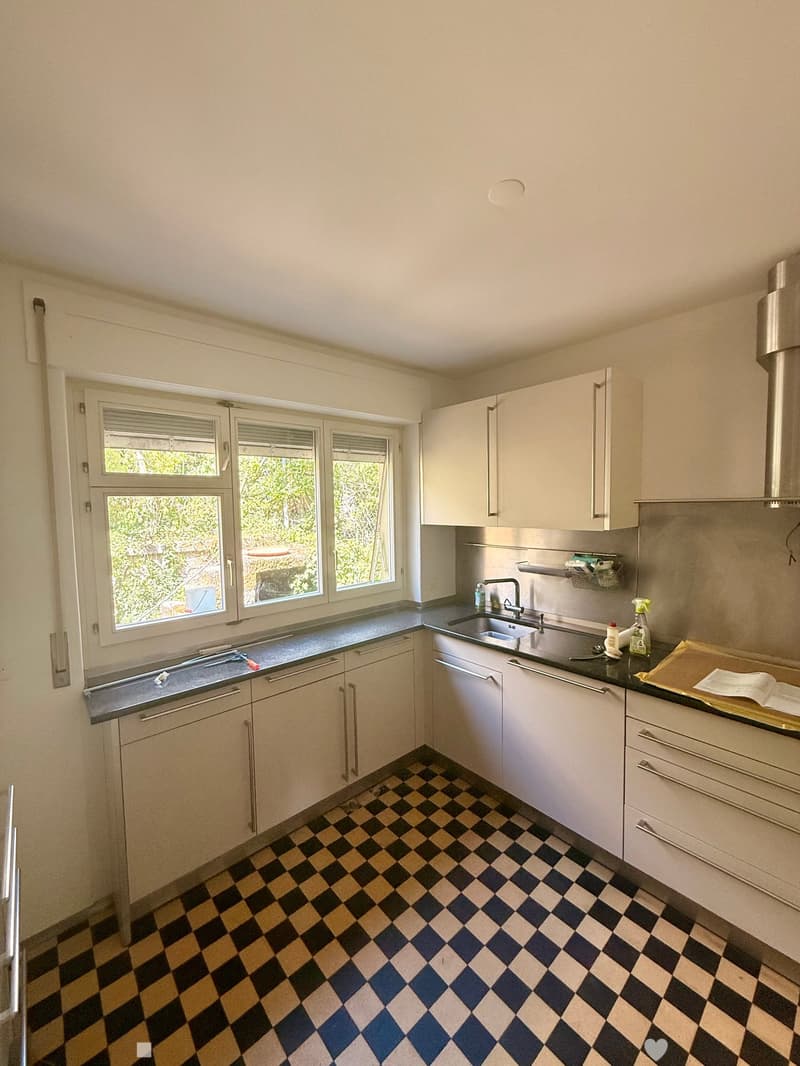 Modern kitchen with white cabinets, black countertop, checkered floor, and large windows overlooking greenery.