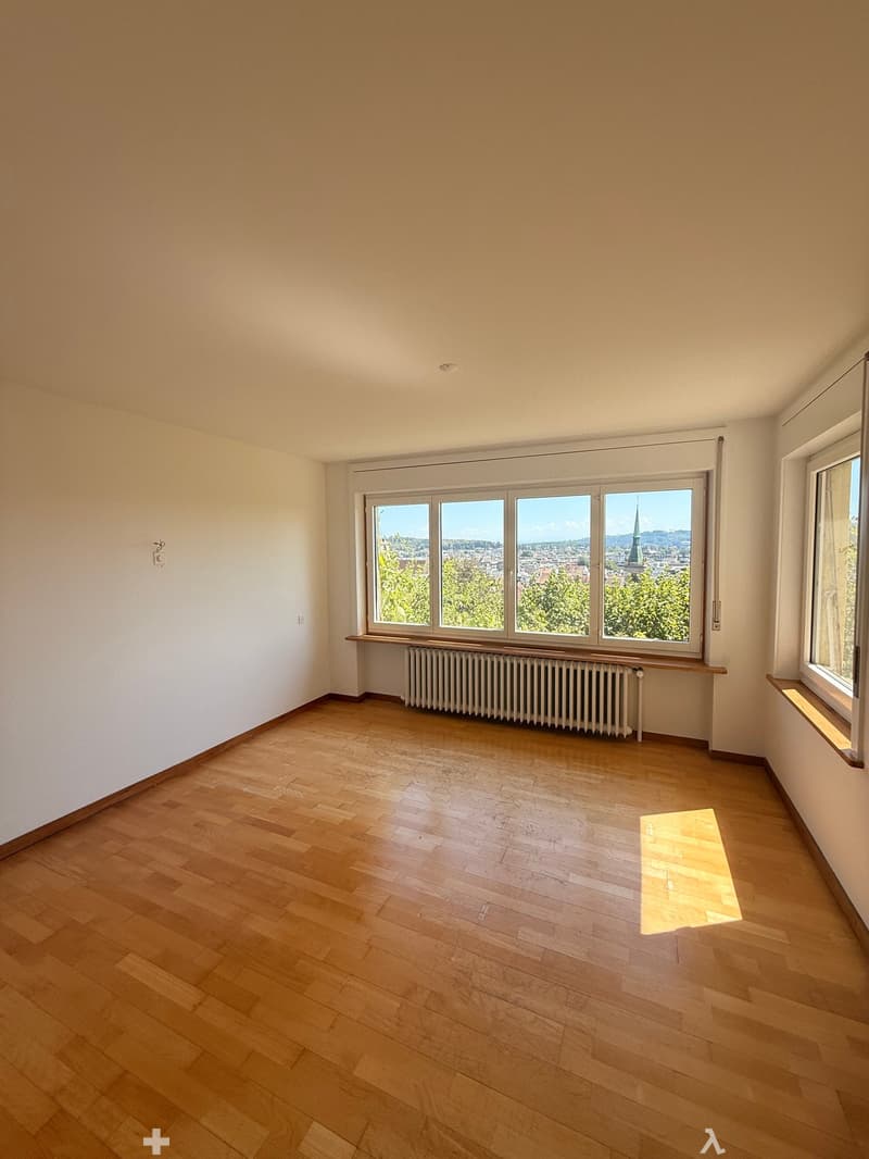Empty room with wooden flooring, large windows offering a cityscape view, radiator, and natural light.