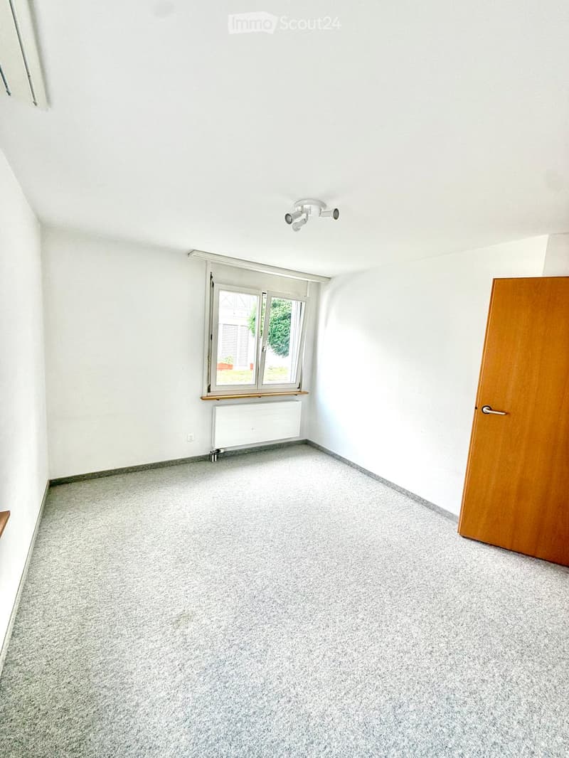 Empty room with white walls, grey carpeted floor, large window with blinds, radiator, and wooden door.