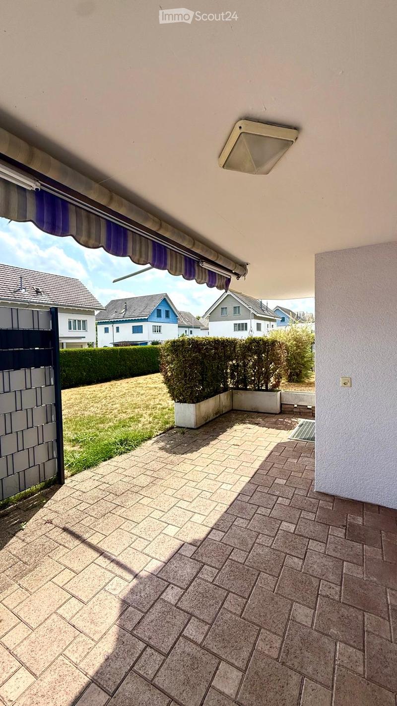 Balcony with tiled floor, covered by a retractable awning, overlooking a well-maintained garden and neighboring houses.