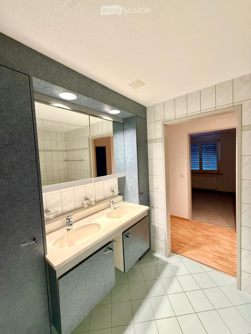 Modern bathroom with double sink, large mirror, and built-in cabinets. White tiled floor and walls, with a view into an adjacent room with wooden flooring.