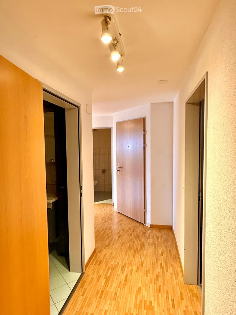 A narrow hallway with wooden flooring, white walls, and several doors leading to different rooms. The hallway is illuminated by ceiling lights.