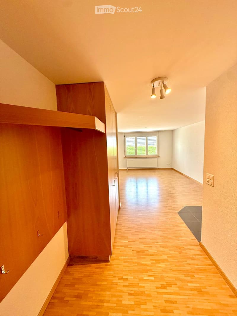 Empty hallway with wooden flooring, white walls, and a ceiling light fixture. A closed wooden door is visible on the left, and a window with blinds is at the end of the hallway.