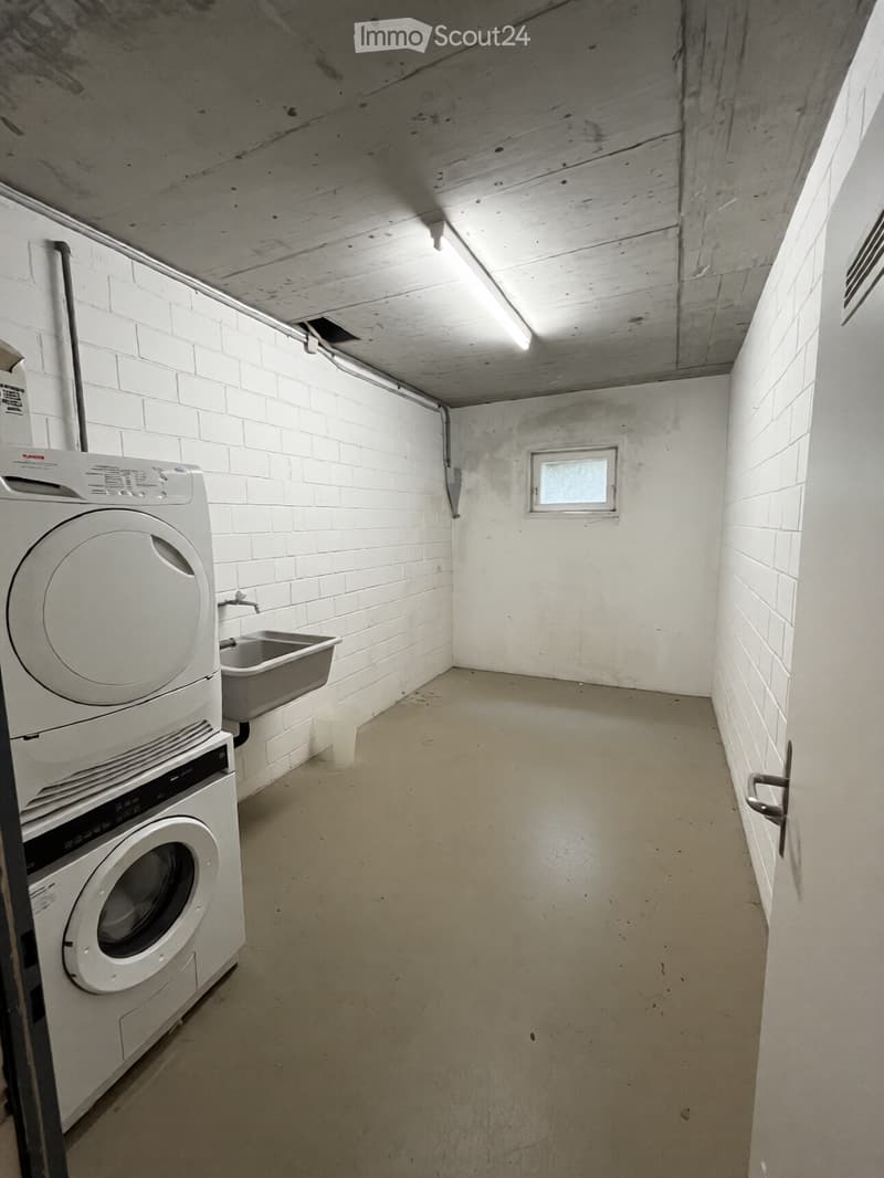Utility room with a washing machine, sink, and a small window. The walls are white brick, and the floor is concrete.