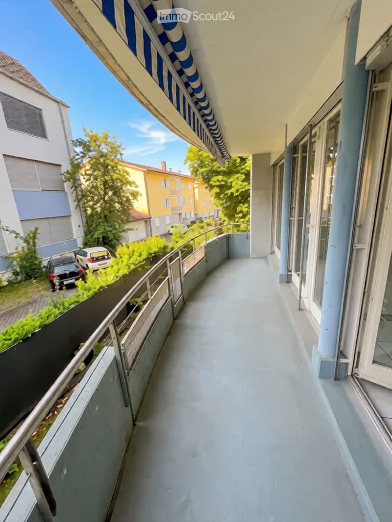 Balcony with a striped awning, metal railing, and a view of neighboring buildings and parked cars.