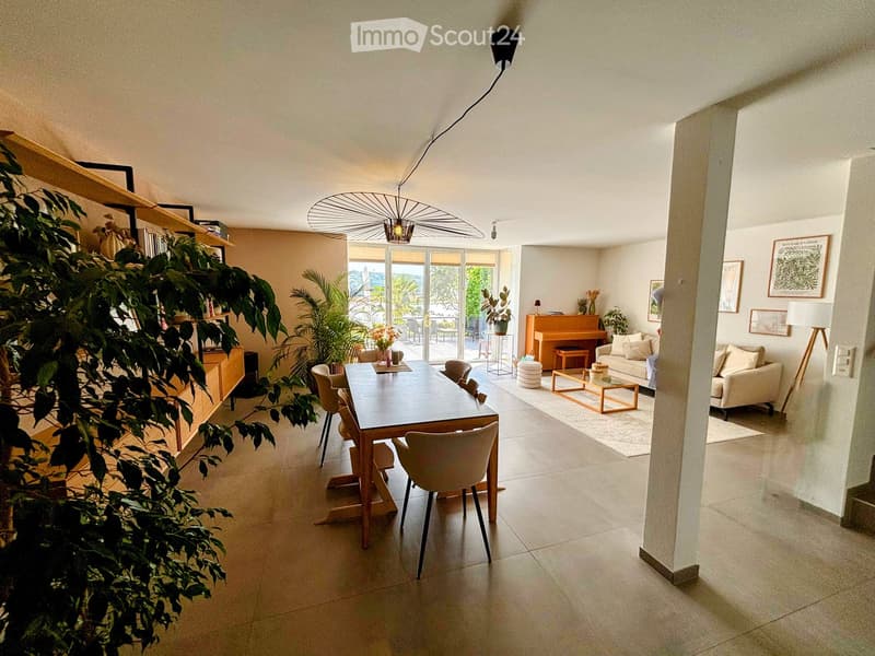Open-plan living and dining area with a sofa, coffee table, piano, dining table with chairs, bookshelf, and decorative plants.