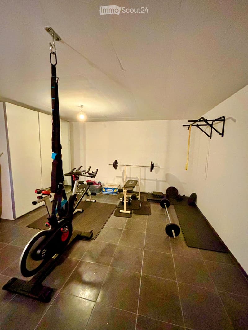 A small, well-lit room with tiled flooring and white walls, containing various exercise equipment including an exercise bike, a barbell, a weight bench, dumbbells, and yoga mats.