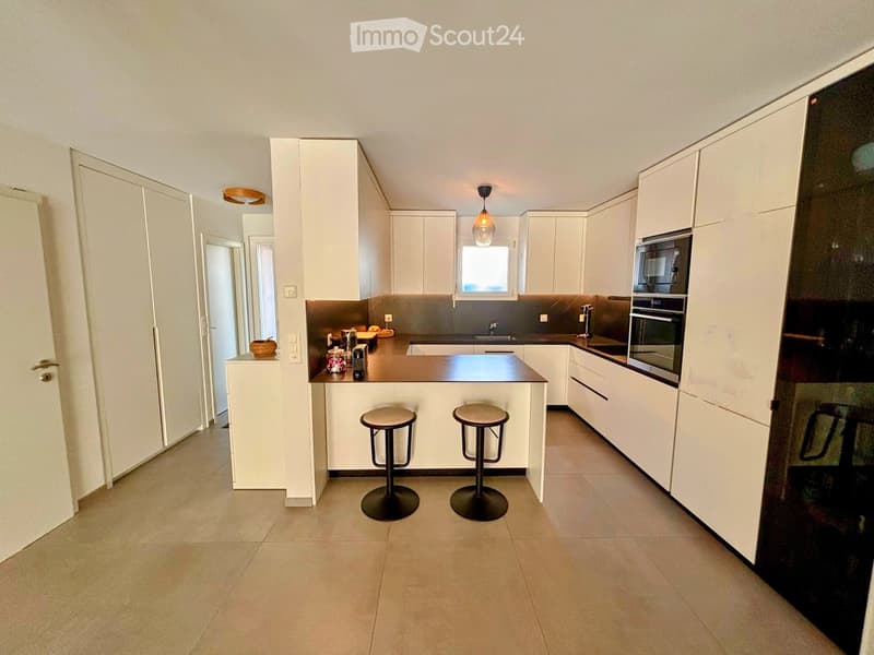 Modern kitchen with white cabinets, dark countertops, and built-in appliances. Two stools are placed at the island, and a small window provides natural light.